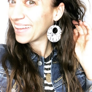 May include: A woman with long brown hair is wearing a denim jacket over a black and white striped shirt. She is wearing large, round, white and black patterned earrings.