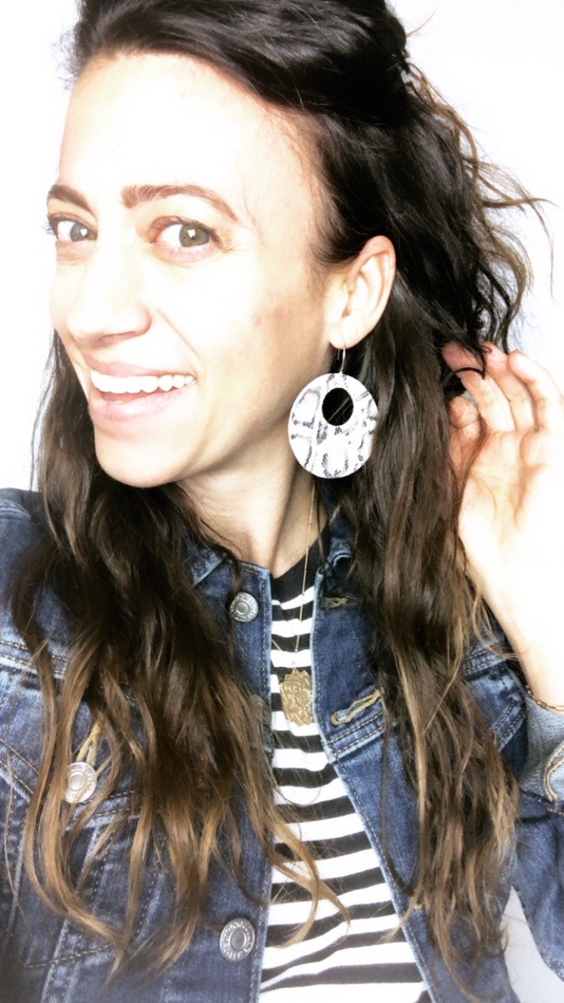 May include: A woman with long brown hair is wearing a denim jacket over a black and white striped shirt. She is wearing large, round, white and black patterned earrings.