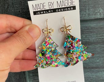 Puede incluir: Un par de pendientes de árbol de Navidad con lazos y ganchos dorados. Los pendientes están hechos de material transparente lleno de purpurina de colores. Se muestran en una tarjeta blanca con el texto "MADE BY MAGGIE JEWELRY DESIGNS".