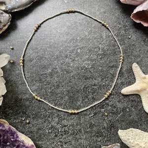 K&ouml;nnte beinhalten: Zarte silberne Halskette mit gleichm&auml;&szlig;ig verteilten goldenen Perlen. Die Halskette ist kreisf&ouml;rmig und liegt auf einer dunklen, strukturierten Oberfl&auml;che. Der Hintergrund umfasst Kristalle, Muscheln und einen Seestern.