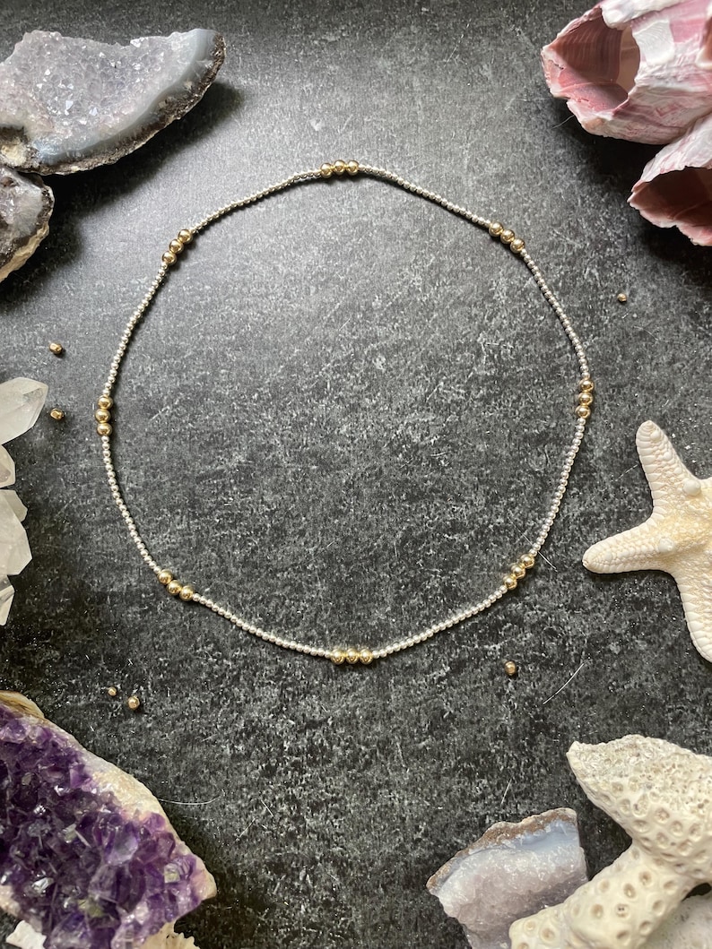 K&ouml;nnte beinhalten: Zarte silberne Halskette mit gleichm&auml;&szlig;ig verteilten goldenen Perlen. Die Halskette ist kreisf&ouml;rmig und liegt auf einer dunklen, strukturierten Oberfl&auml;che. Der Hintergrund umfasst Kristalle, Muscheln und einen Seestern.