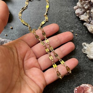 May include: A gold chain necklace with a flat oval link design. The chain is shown on a hand against a dark background.
