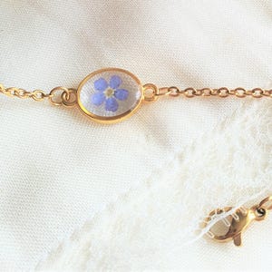 May include: Gold-toned chain bracelet featuring a small, oval pendant with a delicate blue flower encased in clear resin. The bracelet has a lobster clasp closure. The background is a white, textured fabric.