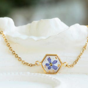 May include: A gold chain bracelet with a hexagonal pendant featuring a pressed purple flower encased in clear resin.