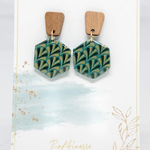Puede incluir: Pendientes con parte superior de madera y colgantes hexagonales en color turquesa y dorado. Los pendientes se exhiben en una tarjeta blanca con el texto "inspired by the beauty of nature" y el nombre de la tienda "Raffinesse".
