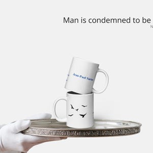 May include: Two white ceramic mugs stacked on a silver tray. The top mug has the text "Jean-Paul Sartre" in blue. The bottom mug features black bird silhouettes. The phrase "Man is condemned to be free" is above the mugs.