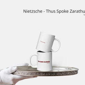 Puede incluir: Dos tazas de cerámica blanca apiladas sobre una bandeja plateada, sostenidas por una mano enguantada. La taza superior tiene la palabra "Derniersch" impresa en rojo. La taza inferior tiene el texto "Thus Spoke Zarathustra" impreso en rojo.