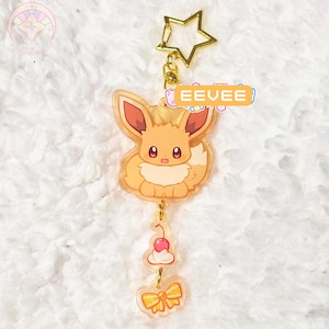May include: A clear acrylic keychain featuring a cartoon Eevee character with a gold star-shaped clasp. The keychain includes a cherry and bow charm. The word "EEVEE" is printed on a rectangular tag.