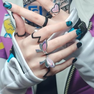 May include: A close-up of hands adorned with rings and nail art. The rings feature heart and floral designs, with black and silver accents. The nails are painted black and teal with glitter.