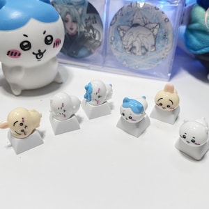 Peut inclure: Ensemble de touches de clavier décoratives avec des personnages de dessins animés. Les touches sont blanches avec des visages détaillés et des accents bleus, roses et jaunes. D'autres objets de collection sont également visibles.