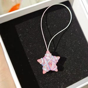 May include: A pink star-shaped charm with a white and pink string attached. The charm is in a white box with a black interior.