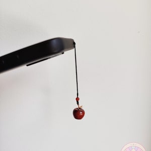 May include: A small, red apple charm with a black cord and red bead accents. The charm is suspended from a black object, possibly a phone. The shop name "ioniatrinketshop" is printed in black at the top of the image.