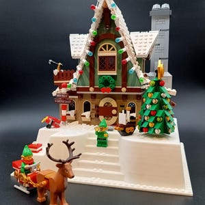 Pode incluir: Um conjunto Lego com tema natalino apresentando uma oficina com uma placa que diz "North Pole Workshop". O conjunto inclui uma árvore de Natal, elfos, uma rena puxando um trenó e luzes decorativas. O edifício tem uma fachada verde e marrom.