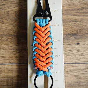 以下が含まれることがあります： 黒い金属製のクリップとリングが付いた、手作りのパラコードキーホルダー。キーホルダーは、鮮やかなオレンジとターコイズブルーの織り模様が特徴です。キーホルダーの長さは約15cmです。