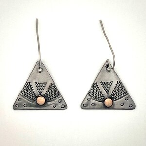 Puede incluir: Un par de pendientes triangulares con acabado en plata oxidada oscura, un patr&oacute;n de puntos texturizado y una peque&ntilde;a cuenta redonda de color cobre centrada en la base de cada tri&aacute;ngulo, suspendidos de sencillos ganchos de color plateado.
