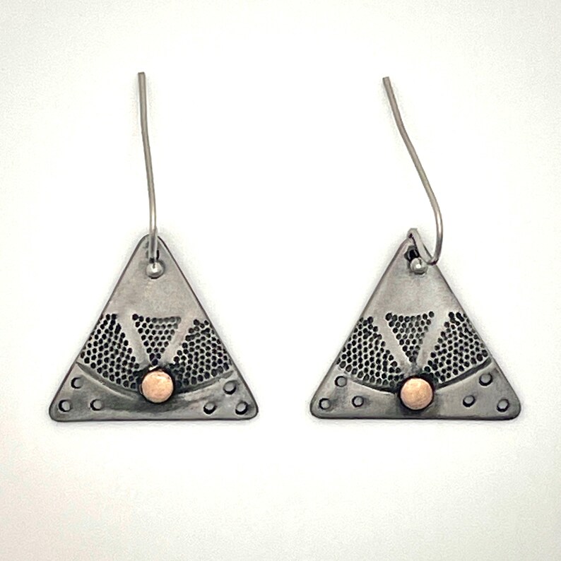 Puede incluir: Un par de pendientes triangulares con acabado en plata oxidada oscura, un patr&oacute;n de puntos texturizado y una peque&ntilde;a cuenta redonda de color cobre centrada en la base de cada tri&aacute;ngulo, suspendidos de sencillos ganchos de color plateado.