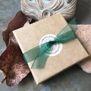 Puede incluir: Una caja de regalo cuadrada de cart&oacute;n marr&oacute;n claro atada con una cinta transparente de color verde oscuro. Una pegatina blanca circular est&aacute; centrada en la tapa, con el texto "GILL GIBSON JEWELRY" y "gillgibsonjewelry.etsy.com" en letras negras.