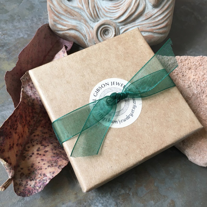 Puede incluir: Una caja de regalo cuadrada de cart&oacute;n marr&oacute;n claro atada con una cinta transparente de color verde oscuro. Una pegatina blanca circular est&aacute; centrada en la tapa, con el texto "GILL GIBSON JEWELRY" y "gillgibsonjewelry.etsy.com" en letras negras.