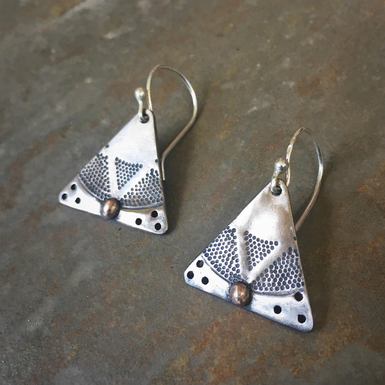 Puede incluir: Un par de pendientes triangulares de plata oxidada, con un patr&oacute;n de textura punteada y una peque&ntilde;a cuenta de color cobre en el centro. Cada pendiente cuelga de un gancho de plata curvado y presenta un acabado oscuro y desgastado.
