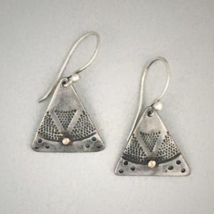 Puede incluir: Un par de pendientes triangulares colgantes de plata oxidada, con un patr&oacute;n de puntos texturizado y una peque&ntilde;a cuenta dorada en el centro. Cada pendiente cuelga de un gancho de plata curvado y presenta un acabado oscuro y desgastado.
