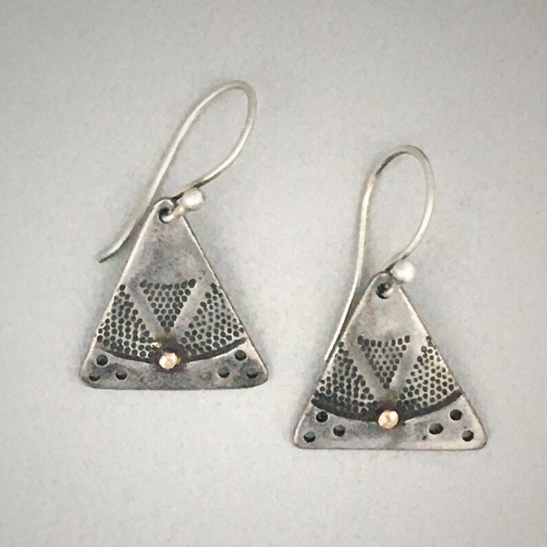 Puede incluir: Un par de pendientes triangulares colgantes de plata oxidada, con un patr&oacute;n de puntos texturizado y una peque&ntilde;a cuenta dorada en el centro. Cada pendiente cuelga de un gancho de plata curvado y presenta un acabado oscuro y desgastado.