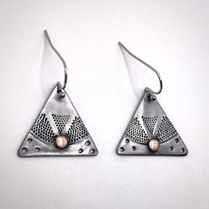 Puede incluir: Un par de pendientes colgantes triangulares hechos de metal plateado oscuro oxidado. Cada pendiente presenta un patr&oacute;n texturizado en forma de abanico de peque&ntilde;os puntos, un peque&ntilde;o acento circular color cobre en la base y ganchos de plata.