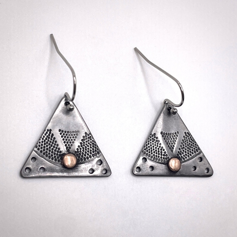 Puede incluir: Un par de pendientes colgantes triangulares hechos de metal plateado oscuro oxidado. Cada pendiente presenta un patr&oacute;n texturizado en forma de abanico de peque&ntilde;os puntos, un peque&ntilde;o acento circular color cobre en la base y ganchos de plata.