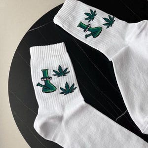 Könnte beinhalten: Paar weiße Socken mit grünen Cannabisblatt- und Bong-Grafiken. Die Socken bestehen aus einem weichen Material und sind auf Komfort ausgelegt. Das Design ist eine lustige und einzigartige Möglichkeit, sich auszudrücken.