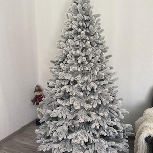 Può includere: Un grande albero di Natale artificiale con un aspetto innevato e floccato. L'albero è di colore grigio chiaro e ha una forma piena e realistica. L'albero si trova in un angolo di una stanza.