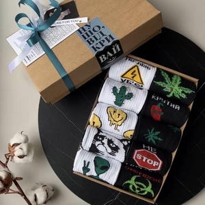 Op de afbeelding: Een geschenkdoos met een set sokken. De sokken hebben verschillende ontwerpen, waaronder een cactus, een smeltend smiley-gezicht, een kattengezicht en een alien. De doos is vastgebonden met een blauwgroen en wit lint.