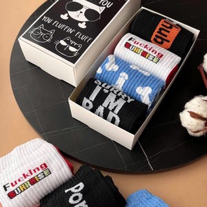 Könnte beinhalten: Ein Geschenkbox-Set mit Novelty-Socken. Der Deckel der Box zeigt Katzenillustrationen und den Text "FLUFF YOU" und "YOU FLUFFIN' FLUFF". Die Socken sind schwarz, weiß und blau, mit verschiedenen grafischen Designs und Texten, darunter "hub", "FUCKING SURPRISE" und "SORRY I'M BAD".