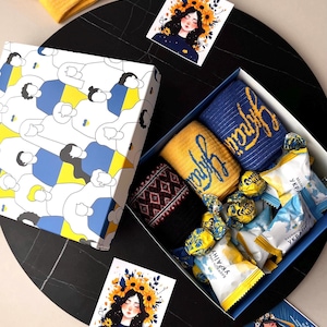 May include: A gift box featuring Ukrainian themed items. The box contains socks with the word "Ukraine" in yellow script, chocolates, and cards with illustrations of women with floral crowns. The box has a blue and yellow design.