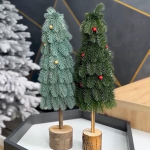 Op de afbeelding: Twee miniatuur kunstkerstbomen. Eén is blauwgroen met gouden ornamenten, de andere is groen met rode ornamenten. Beide bomen zijn gemonteerd op houten voetstukken en worden tentoongesteld op een wit oppervlak.