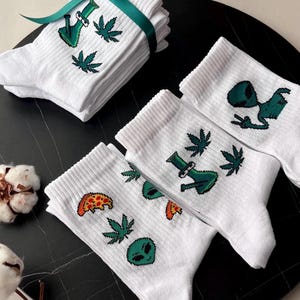 Può includere: Calzini bianchi con grafiche verdi di foglie di cannabis, alieni, pizza e bong. Una pila di calzini è legata con un nastro verde. I calzini sono su una superficie nera.