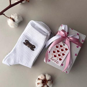 Può includere: Calzini bianchi con un disegno di orsetto marrone, accanto a una confezione regalo avvolta in carta fantasia e legata con un nastro rosa. Un'etichetta con cuori rossi e la parola "love" è attaccata. Sono presenti anche rametti di cotone.