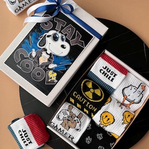Puede incluir: Un juego de calcetines de regalo con varios diseños. La caja presenta una ilustración de Snoopy con el texto "STAY COOL". Los calcetines incluyen diseños con caras sonrientes, un dedo medio y las palabras "JUST CHILL" y "GAMER".