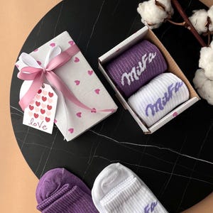 May include: A gift set featuring socks, a gift box, and a tag. The socks are purple and white with the word "Mira" in a script font. The gift box is white with pink hearts and a pink ribbon. A tag says "love".
