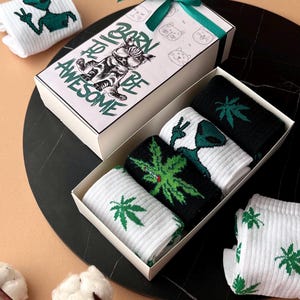 Pode incluir: Um conjunto de caixa de presente de meias com vários designs. A caixa apresenta um gráfico de gato e o texto "Born to be Awesome". As meias incluem padrões de folhas alienígenas e de cannabis em verde e preto em meias brancas. Uma fita verde adorna a caixa.