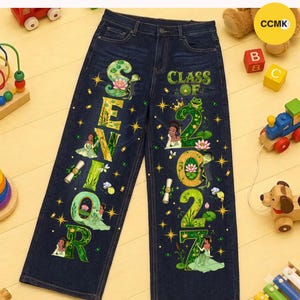May include: Dark blue denim jeans featuring a graphic print with the text "SENIOR" and "CLASS OF 2027". The design includes illustrations of a character in a green dress, a frog wearing a crown, lotus flowers, graduation caps, and scrolls.