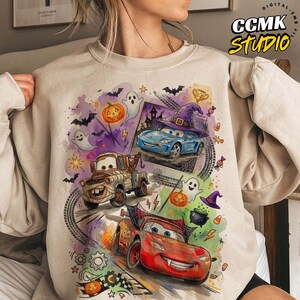 May include: A beige crewneck sweatshirt featuring a colorful Halloween-themed graphic print of three animated cars, including a red race car, a brown tow truck, and a blue car wearing a witch hat, surrounded by ghosts, bats, and pumpkins.