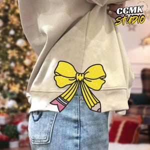 Puede incluir: Sudadera beige con diseño de lazo y lápiz amarillo. El lazo está delineado en negro y tiene un efecto de brillo amarillo. El lápiz tiene una goma de borrar rosa y una punta gris. El texto "CCMK Studio" está en la esquina superior derecha.