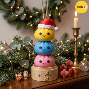 May include: A stack of three round, smiling dumpling ornaments in yellow, blue, and pink, topped with a red and white Santa hat. The stack sits in a small bamboo steamer base, suspended by a twine string, with a yellow CCMK logo in the corner.