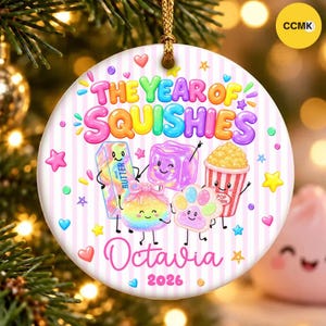 Puede incluir: Un adorno redondo de cerámica blanca con rayas rosas verticales que presenta el texto THE YEAR OF SQUISHIES y Octavia 2026. El diseño incluye ilustraciones coloridas de mantequilla, un cubo, un peluche redondo y palomitas.