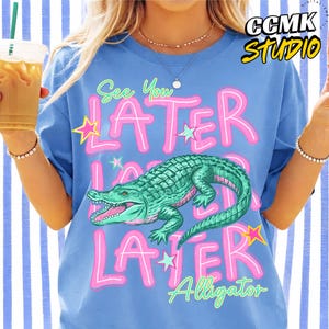 May include: A light blue t-shirt featuring the text "See You Later" in pink with a white outline, and a green alligator illustration. The word "Alligator" is written below the alligator in green. The shirt has a loose fit.