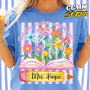 May include: A light blue t-shirt featuring a colorful floral design emerging from an open book. The shirt includes the text "Teach them, love them, watch them grow" and "Mrs. Kaycee" on a pencil graphic. The CCMK Studio logo is in the upper right corner.