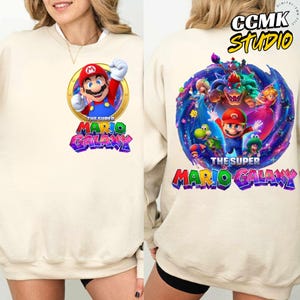 Pode incluir: Camisola creme com um gr&aacute;fico colorido de Mario de "The Super Mario Galaxy". O design frontal mostra Mario num c&iacute;rculo dourado, enquanto a parte de tr&aacute;s apresenta v&aacute;rios personagens. O texto "THE SUPER MARIO GALAXY" &eacute; impresso em ambos os lados.