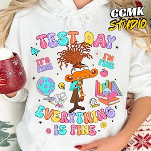 Op de afbeelding: Witte hoodie met een kleurrijke afbeelding met een stripfiguur met de tekst "TEST DAY" en "EVERYTHING IS FINE". Het ontwerp bevat schoolthema-elementen zoals boeken, een wereldbol en een appel.