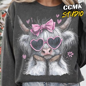 May include: Dark gray sweatshirt featuring a whimsical illustration of a Highland cow. The cow wears heart-shaped sunglasses and a pink bow. The design includes pink hearts and a flower, with the text "CCMK STUDIO" in yellow.