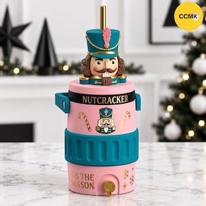 May include: A pink and teal insulated tumbler featuring a nutcracker figure on the lid with a gold straw. The body displays the text NUTCRACKER and TIS THE SEASON with gold candy cane and snowflake graphics. It has a teal textured grip band.