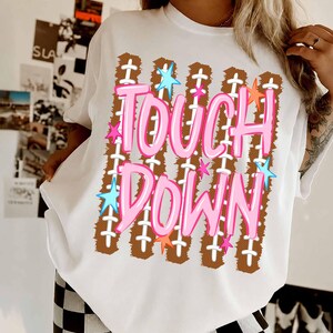 May include: A white t-shirt featuring a graphic print of brown football-style textured stripes with white laces. The text TOUCH DOWN is printed in large, bright pink bubble letters, accented by small, colorful blue, pink, and orange stars.